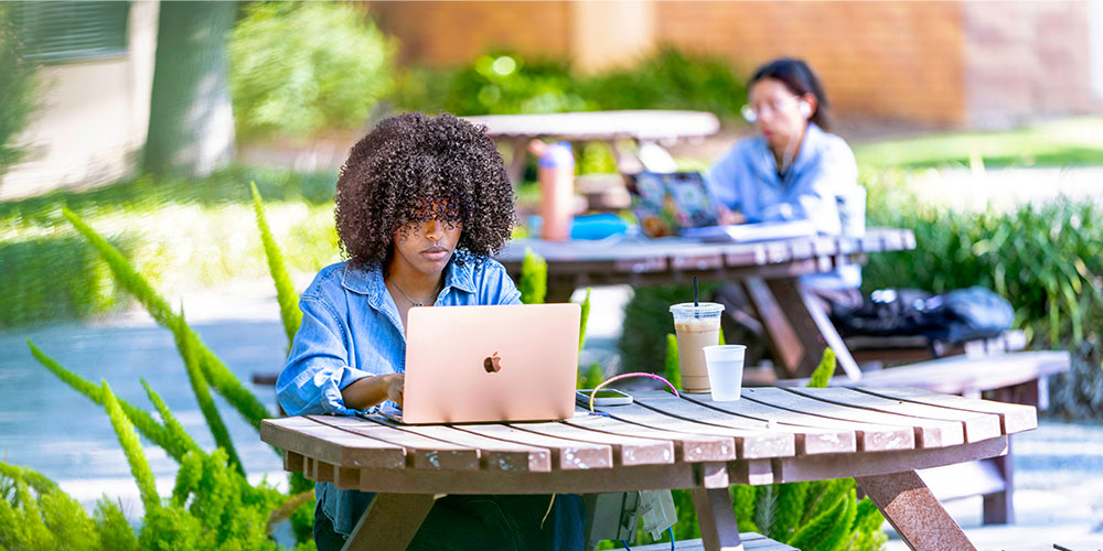 A Fresno Pacific University student works on a laptop at a picnic table on campus.