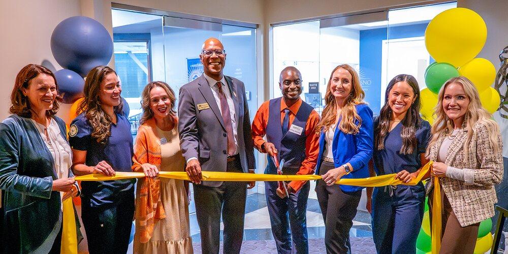 Eight Fresno Pacific University and CalViva Health representatives gather with scissors and a yellow ribbon during a ribbon-cutting ceremony.