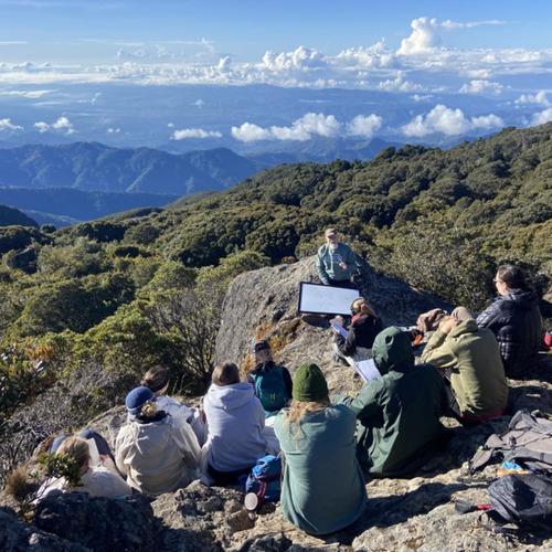 Students sit on a rocky mountain overlook during an outdoor lesson above forested valleys in Costa Rica.