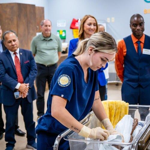 Nursing student in scrubs cares for an infant mannequin in a bassinet while faculty and guests observe.