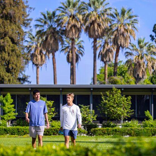 Two Fresno Pacific University students walk across a sunny campus lawn with palm trees and a campus building in the background.