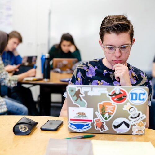 An FPU student concentrates on an assignment using a sticker-covered laptop in a study room.