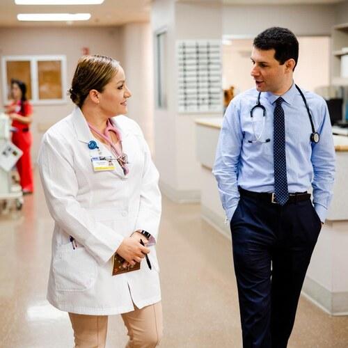 Two health care professionals wearing stethoscopes talk while walking through a hospital hallway.