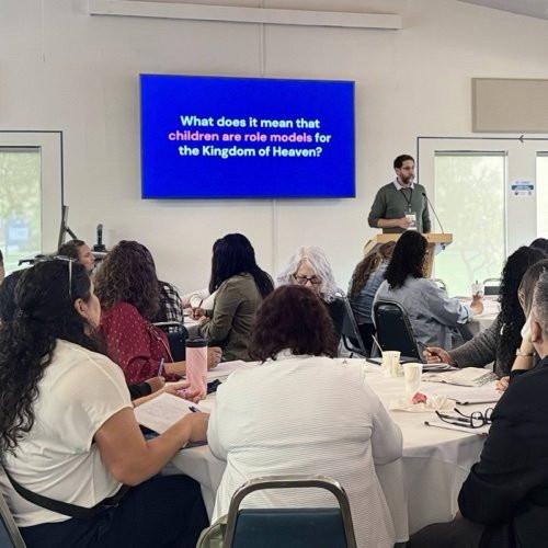 Children's ministry leaders seated at round tables listen to a presenter beside a screen asking what it means that children are role models for the Kingdom of Heaven