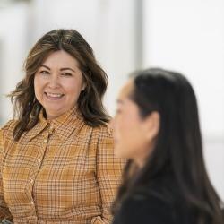 Two women talk together in a bright room, with one smiling toward the camera.