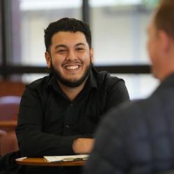 A smiling FPU student talks with another person while seated at a table in a campus lounge.