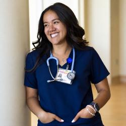 Smiling nursing student in navy scrubs poses with a stethoscope in a campus hallway.