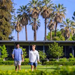 Two Fresno Pacific University students walk across a sunny campus lawn with palm trees and a campus building in the background.