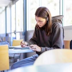 An FPU student wearing earbuds writes in a notebook beside large windows in a campus study space.