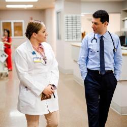 Two health care professionals wearing stethoscopes talk while walking through a hospital hallway.